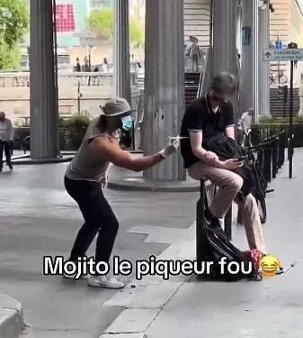 Una persona con un sombrero, cara facial y guantes que fingen inyectar a una persona sentada en un banco con una jeringa.