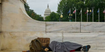 Una persona sin hogar duerme en un banco con el edificio del Capitolio de Estados Unidos al fondo.