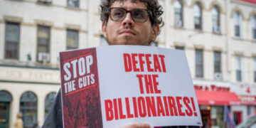 Un manifestante sostiene un cartel que dice "DETENGAN LOS RECORTES; DERROTAR A LOS MULTIMILLONARIOS" mientras participaba en una manifestación al aire libre