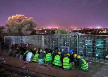 Trabajadores migrantes con chalecos amarillos brillantes se apiñan en el suelo mientras un brillante horizonte ilumina el cielo detrás de ellos.
