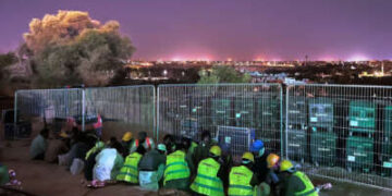 Trabajadores migrantes con chalecos amarillos brillantes se apiñan en el suelo mientras un brillante horizonte ilumina el cielo detrás de ellos.