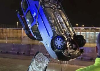 Un collage de imágenes que contiene 1 imágenes. La imagen 1 muestra un automóvil azul y blanco que una grúa levanta boca abajo desde el agua por la noche, con una persona con un chaleco de seguridad mirando