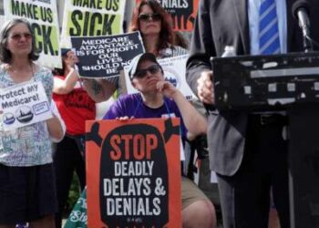 Un manifestante sosteniendo un cartel que decía "DETENGA RETRASOS Y NEGACIONES MORTALES" durante un mitin al aire libre escucha a alguien con traje hablando frente a un micrófono en primer plano