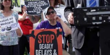 Un manifestante sosteniendo un cartel que decía "DETENGA RETRASOS Y NEGACIONES MORTALES" durante un mitin al aire libre escucha a alguien con traje hablando frente a un micrófono en primer plano