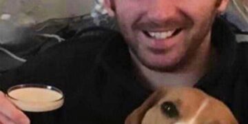 Hombre sonriendo y sosteniendo una pinta de cerveza con un beagle a su lado.