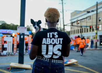 Una mujer negra que sostiene un perro increíblemente lindo y pequeño mira hacia afuera de la cámara y hacia una protesta al aire libre, mientras usa una camiseta que lee "SE TRATA DE NOSOTROS"