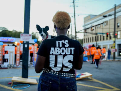 Una mujer negra que sostiene un perro increíblemente lindo y pequeño mira hacia afuera de la cámara y hacia una protesta al aire libre, mientras usa una camiseta que lee "SE TRATA DE NOSOTROS"