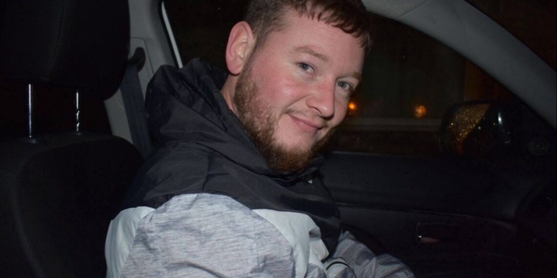 Un hombre con barba y pelo corto sentado en un coche, sonriendo.