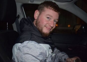Un hombre con barba y pelo corto sentado en un coche, sonriendo.