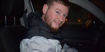 Un hombre con barba y pelo corto sentado en un coche, sonriendo.