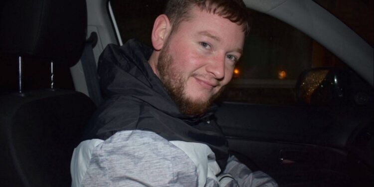 Un hombre con barba y pelo corto sentado en un coche, sonriendo.