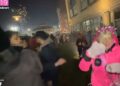 Una mujer con una chaqueta rosa y una corona festiva es atacada por un fuego artificial durante la cuenta regresiva de Año Nuevo.