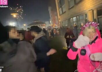 Una mujer con una chaqueta rosa y una corona festiva es atacada por un fuego artificial durante la cuenta regresiva de Año Nuevo.