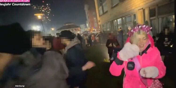 Una mujer con una chaqueta rosa y una corona festiva es atacada por un fuego artificial durante la cuenta regresiva de Año Nuevo.
