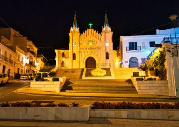 Un collage de imágenes que contiene 1 imágenes. La imagen 1 muestra la vista nocturna de la Capilla de Santa Vera Cruz en Alhaurín El Grande, provincia de Málaga, España, que presenta una torre única de tres secciones y una estructura triangular.