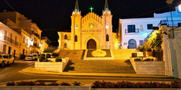 Un collage de imágenes que contiene 1 imágenes. La imagen 1 muestra la vista nocturna de la Capilla de Santa Vera Cruz en Alhaurín El Grande, provincia de Málaga, España, que presenta una torre única de tres secciones y una estructura triangular.
