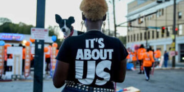 Una mujer negra que sostiene un perro increíblemente lindo y pequeño mira hacia afuera de la cámara y hacia una protesta al aire libre, mientras usa una camiseta que lee "SE TRATA DE NOSOTROS"