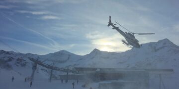 A "Polizei" Helicóptero volando sobre una cadena montañosa nevada con un remonte y un edificio debajo.