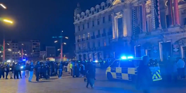 Coches de policía con luces azules intermitentes por la noche frente a un edificio, con gente reunida en primer plano.