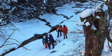 Cuatro personas con ropa de invierno y cascos navegando por un paisaje cubierto de nieve junto a un arroyo helado.