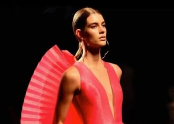 Un collage de imágenes que contiene 1 imágenes. La imagen 1 muestra una modelo con cabello castaño claro, que lleva un vestido rosa brillante con un escote en V pronunciado y un detalle plisado en forma de abanico en los hombros, de pie contra un fondo oscuro.