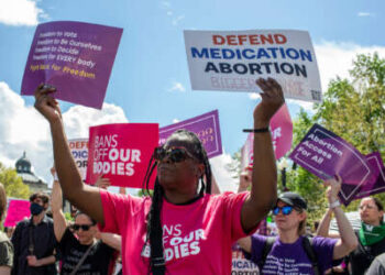 Una mujer negra sostiene carteles que leen "PROTEGER EL ABORTO CON MEDICAMENTOS" y "Libertad de votar; libertad para ser nosotros mismos; libertad para decidir; libertad para TODOS; Luchar por la libertad" durante una protesta al aire libre
